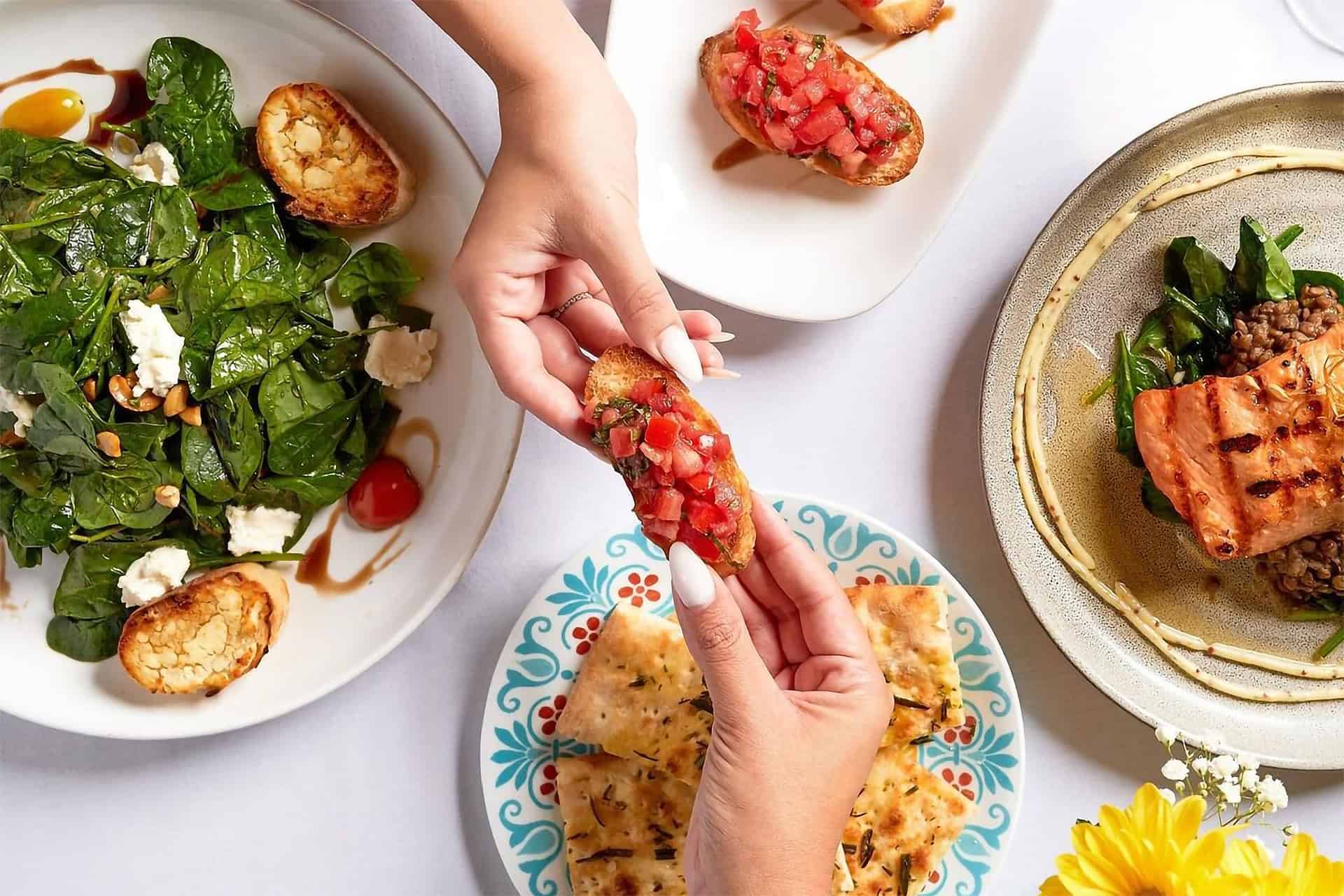 Hands sharing a slice of bruschetta over various other plates of Italian dishes from Serafina restaurant