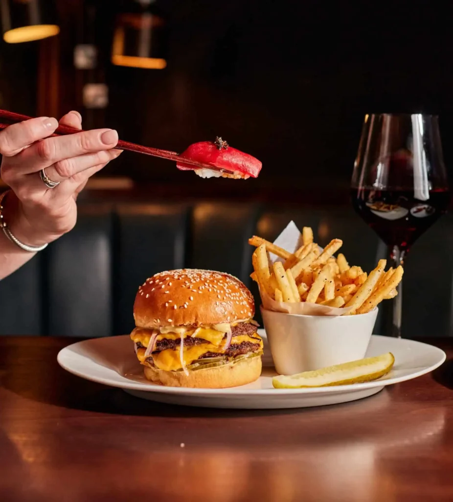 Hand holding a piece of sushi in chopsticks above a plate of burger and fries at Eight Bar